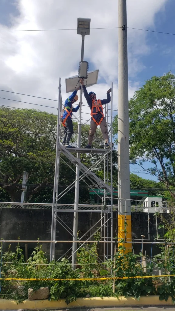 Two NuPON technicians in harnesses on a mobile scaffold installing a solar street light and panel on a roadside pole under a blue sky.