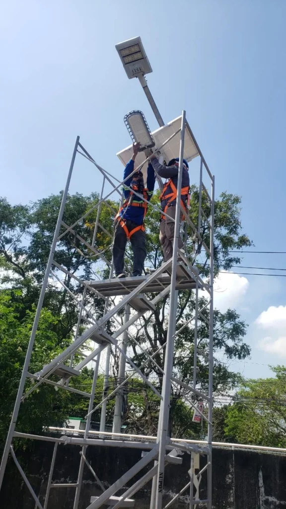 Low-angle view of two harnessed technicians on a scaffold mounting an LED street light beneath a solar panel.