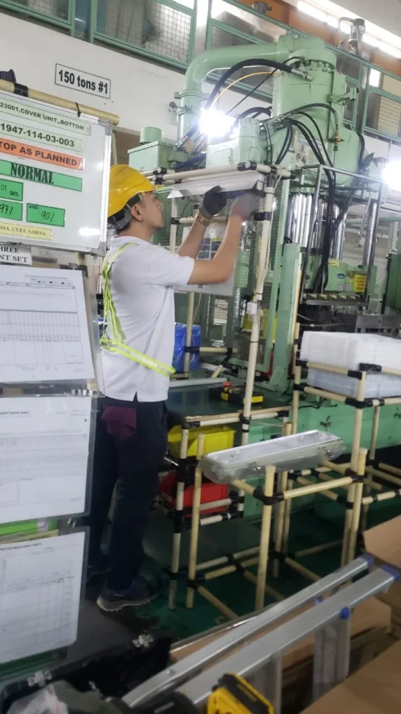 A technician fitting a linear LED fixture beside an injection moulding machine on a production floor.