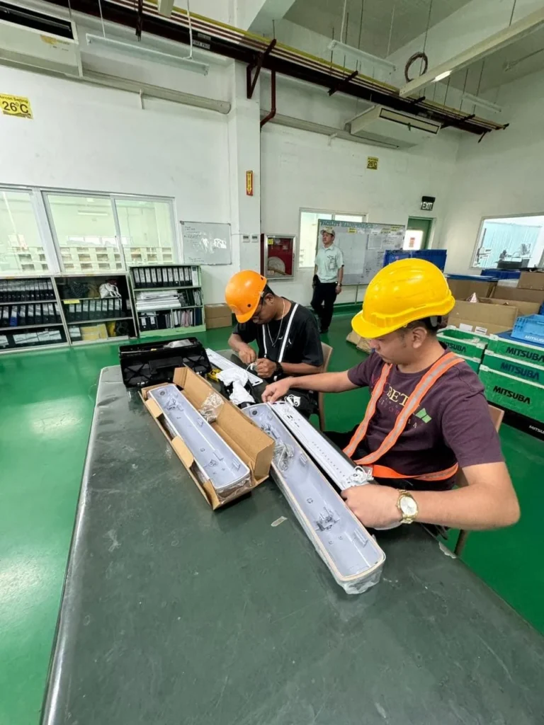Two technicians in hard hats and hi-vis vests assembling LED tri-proof linear fixtures at a workbench.
