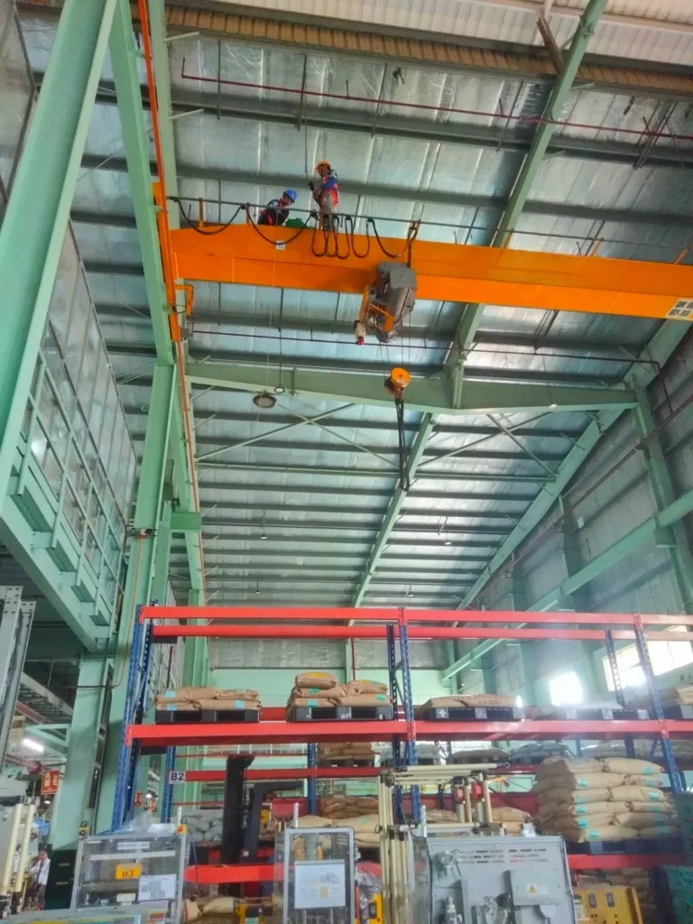 Two technicians working from an orange overhead crane girder high above a factory floor.