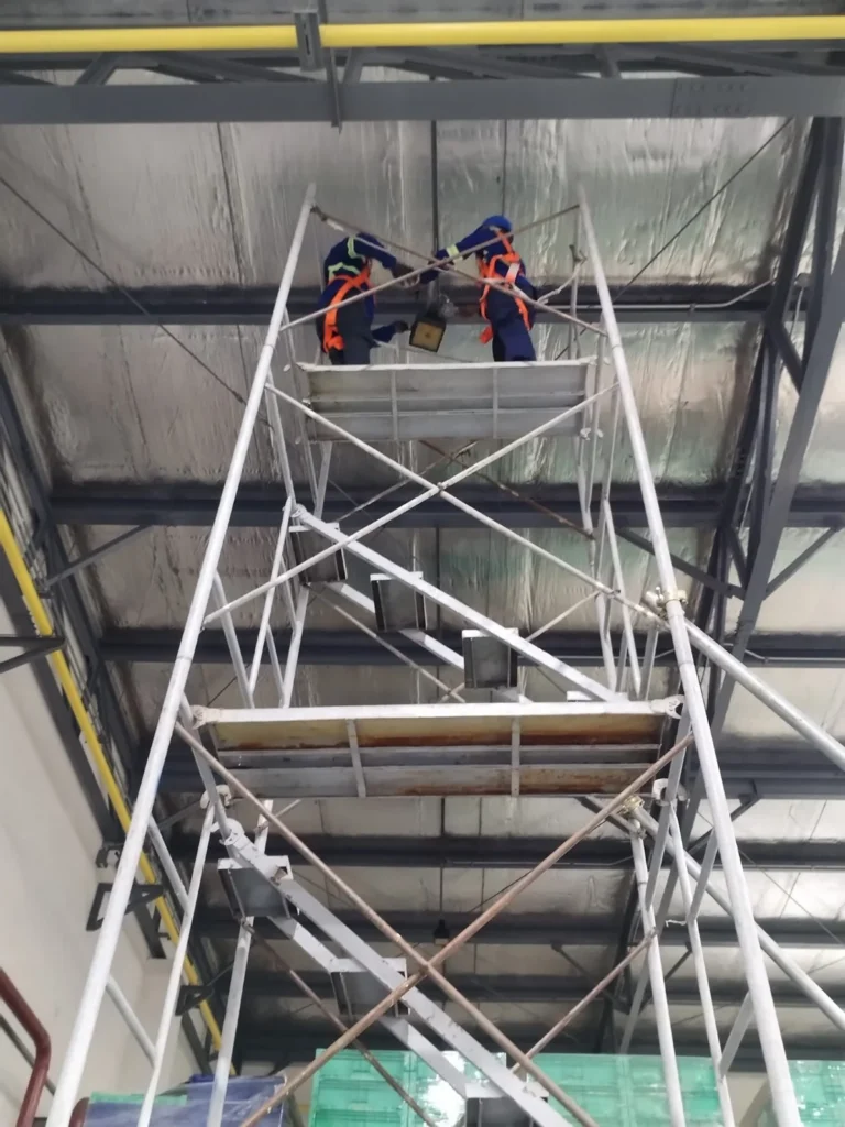 Two technicians in harnesses working at the insulated roof deck of a factory from a tall scaffold tower.
