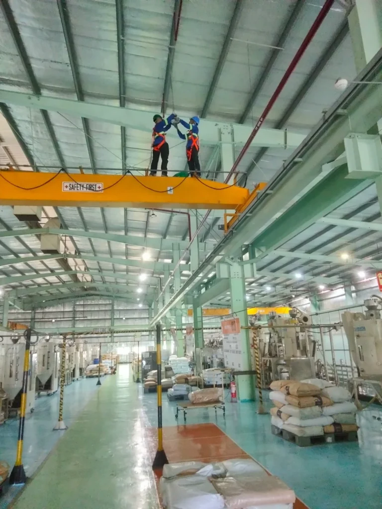 Two harnessed technicians standing on a yellow overhead crane girder to reach ceiling lighting in a production hall.