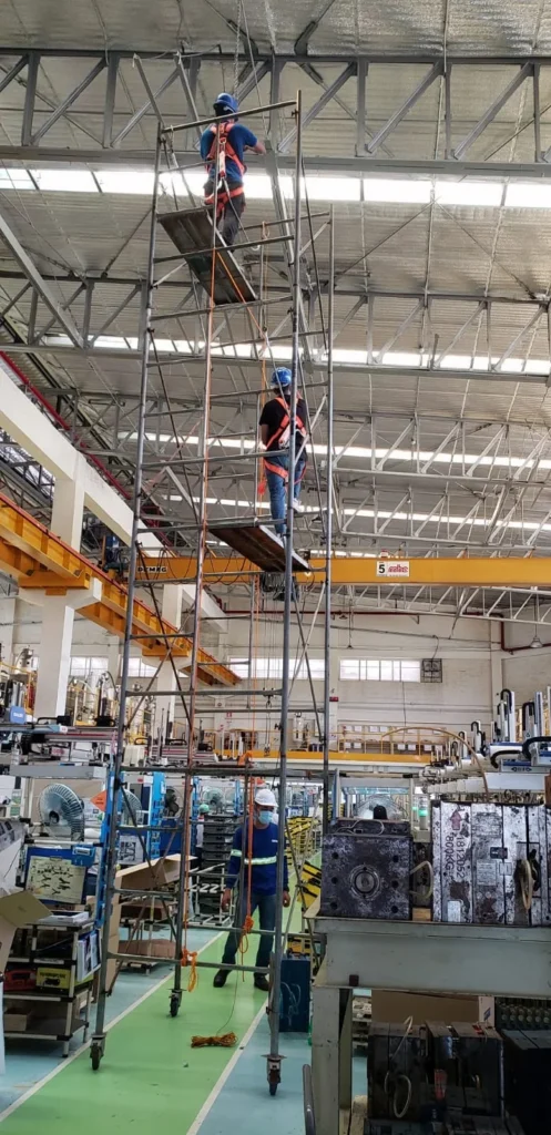 Technicians on a multi-level scaffold tower carrying out lighting work near overhead cranes on a factory floor.